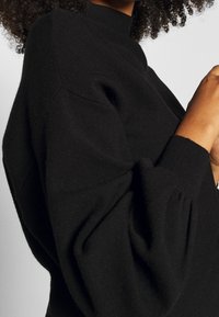 Czarny oversize sweter z wysokim kołnierzem, prążkowanymi mankietami i obszernymi rękawami. Materiał wydaje się być miękki i teksturowany.