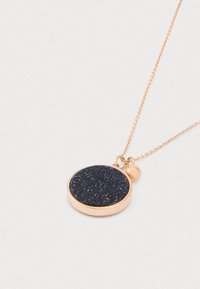 Guld kædethalskæde med rund vedhæng, der har en mørkeblå glitrende sten og en lille guldkugle som charm på hvid baggrund.