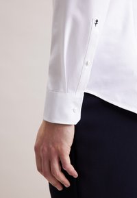 Close-up van de arm van een persoon die een wit overhemd draagt met een kleine geborduurde zwarte roos op de manchet en een donkere broek.