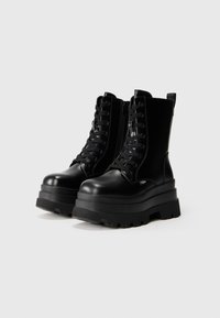 Botas de combate gruesas y negras que cuentan con un upper de cuero suave, cordones en la parte delantera, cremallera lateral y suela de goma gruesa y texturizada.