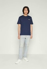 Μπλε ναυτικό βαμβακερό T-shirt με κοντά μανίκια και στρογγυλή λαιμόκοψη, με μικρό λευκό λογότυπο στο αριστερό μέρος του στήθους, συνδυασμένο με ανοιχτόχρωμο τζιν.