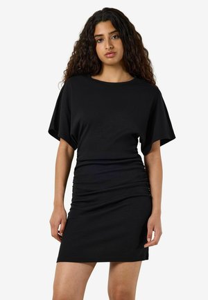 Femme aux longs cheveux bouclés portant une courte robe noire à manches larges, debout devant un fond clair uni.