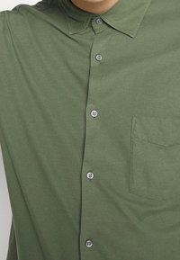 Olivově zelená košilová košile s límečkem, se záplatovou kapsou a hladkou texturou materiálu. Šest knoflíků v kontrastní barvě vpředu.