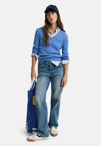 Light blue cable-knit sweater layered over a white collared shirt, paired with light blue jeans and a blue varsity jacket. White sneakers worn.