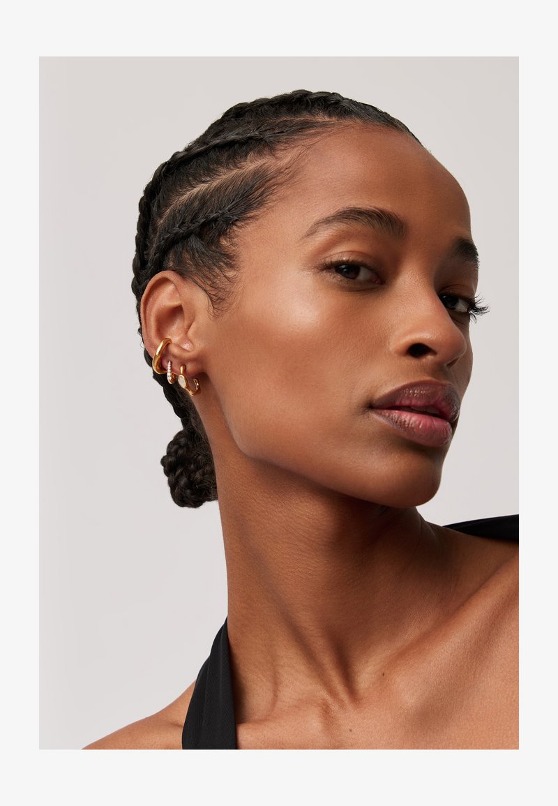 Brincos de argola e de pino em um penteado trançado; pele suave com um brilho natural; vestindo um top halter preto.