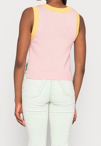 Débardeur en maille côtelée sans manches avec un corps rose et un col jaune. Associé à un pantalon en velours côtelé vert clair avec des rayures verticales et des poches.