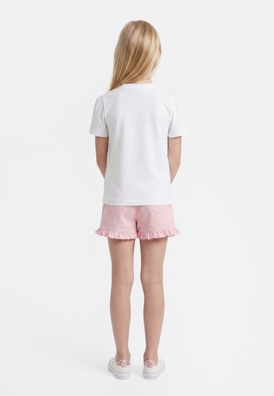 Pikkade blondide juustega tüdruk seisab vastupidiselt, seljas valge lühikeste varrukatega särk, roosad volangidega lühikesed püksid ja valged roosa mustriga tossud.