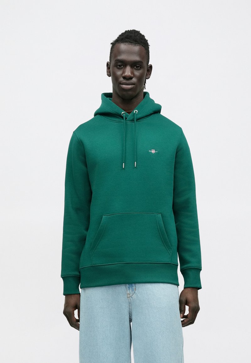 Grøn hættetrøje med en frontlomme, snørrejusterbar hætte og logodetalje på brystet. Kombineret med lyseblå jeans.