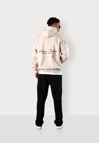 Beige Kapuzenpullover mit schwarzem Drucktext auf dem Rücken, kombiniert mit schwarzen Jogginghosen. Der Kapuzenpullover hat eine große Kapuze und gerippte Bündchen.