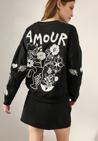 Fekete pulóver fehér virágmintával és az "AMOUR" szóval a hátsó részen. Bordás mandzsettákkal és szegéllyel rendelkezik, fekete szoknyával párosítva.