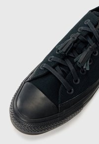 Czarne płócienne trampki z gumowym czubkiem i podeszwą, sznurowane z przodu z czarnymi frędzlami na sznurowadłach, sfotografowane na białym tle.