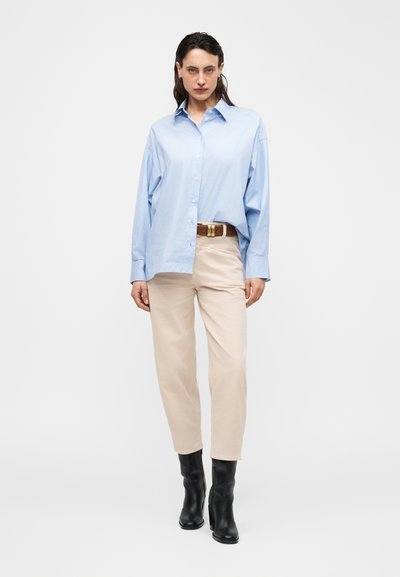 Μία γυναίκα στέκεται φορώντας ανοιχτό γαλάζιο, φαρδύ πουκάμισο με κουμπιά, μπεζ ίσια παντελόνια με καφέ ζώνη και μαύρα μποτάκια μέχρι τον αστράγαλο σε απλό λευκό φόντο.