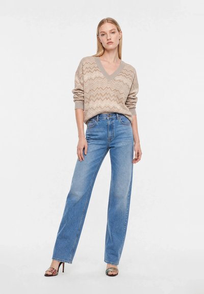 Femme portant un pull beige à motif zigzag, un jean bleu taille haute à jambes larges, et des sandales colorées à talons hauts, debout sur fond blanc.