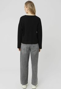 Pull noir à coupe décontractée et ourlet côtelé associé à un jean gris taille haute. Le dos du mannequin est visible sur un fond uni.