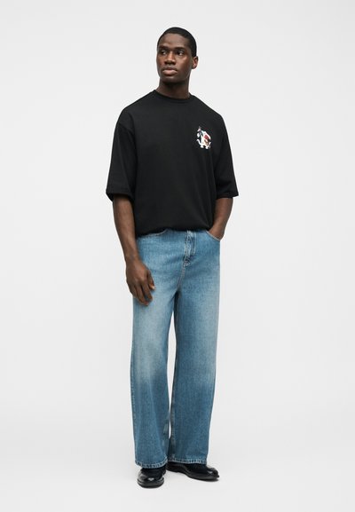 Άνδρας που φοράει μαύρο υπερμέγεθες μπλουζάκι με μικρό σχέδιο, ανοιχτό μπλε τζιν παντελόνι με φαρδιά πόδια, και μαύρα παπούτσια, στέκεται με το ένα χέρι στην τσέπη.