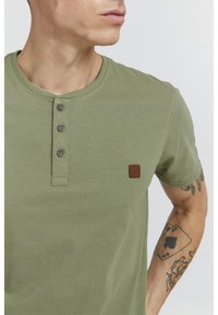 Olive zelené tričko s krátkym rukávom typu henley s trojmočkovou légou, okrúhlym výstrihom a malou hnedou nášivkou na lavom prsníku. Mäkký materiál.