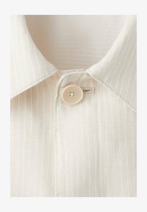 Primer plano de un cuello de camisa beige con textura y un solo botón beige abrochado a través de un ojal.