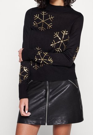 Femme portant un pull noir avec des motifs de flocons de neige dorés et une jupe en cuir noire avec une fermeture éclair à l'avant et des poches zippées.