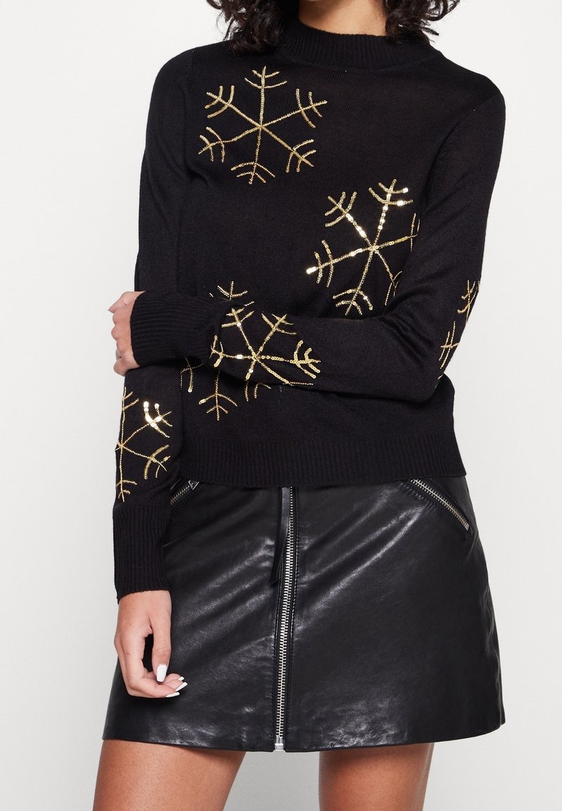 Femme portant un pull noir avec des motifs de flocons de neige dorés et une jupe en cuir noire avec une fermeture éclair à l'avant et des poches zippées.