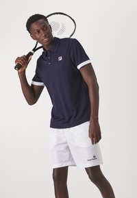 Marineblå polo-skjorte med hvite detaljer, laget av glatt stoff; kombinert med hvite shorts med logo. Tenniskølle holdes over skulderen.