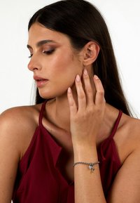 Bracciale in argento con un charm, indossato al polso. Il modello ha i capelli lunghi e scuri, indossa un top bordeaux e posa con le dita che toccano la guancia.
