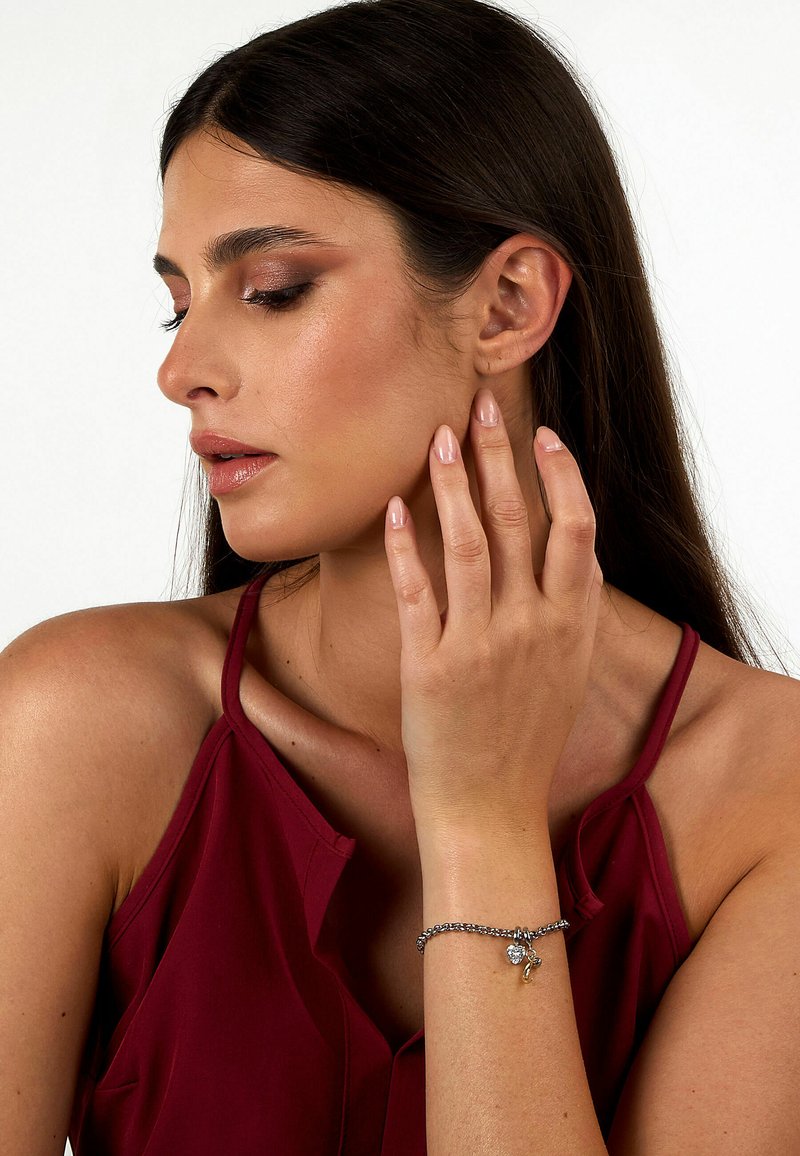 Bracciale in argento con un charm, indossato al polso. Il modello ha i capelli lunghi e scuri, indossa un top bordeaux e posa con le dita che toccano la guancia.