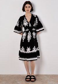 Le mannequin se tient dans une robe noire avec des motifs blancs complexes, présentant un col en V et des manches évasées, associée à des sandales noires.