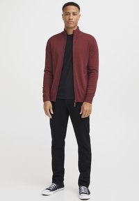 Maglione zip-up color marrone scuro, colletto a costine, indossato su una maglietta nera. Abbinato a jeans neri e sneakers nere con suola bianca.