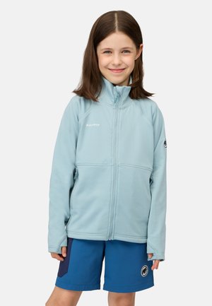Junges Mädchen, das eine hellblaue Jacke mit Reißverschluss und blaue Shorts trägt, lächelt entspannt mit locker hängenden Händen vor einem schlichten Hintergrund.