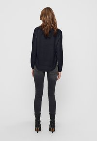 Top blu navy a maniche lunghe con motivo strutturato, scollo rotondo e orlo curvo, abbinato a jeans skinny grigio scuro e stivaletti neri con tacco.