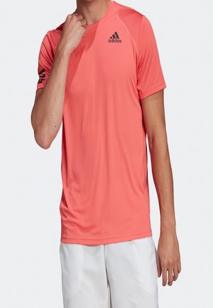 Osoba nosząca koralowy sportowy t-shirt Adidas z krótkim rękawem i czarnym logo na piersi i lewym rękawie, w połączeniu z białymi szortami.