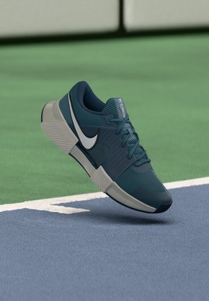 Chaussure de sport Nike bleu sarcelle foncé et blanche flottant au-dessus d'une surface de court de tennis bleu et vert près d'une ligne blanche.
