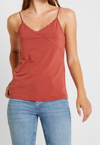 Camisola rosso ruggine con bordi ondulati e spalline regolabili, realizzata in un tessuto liscio e leggero. Abbinata a jeans blu.