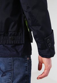 Homme portant une veste bleu marine avec manchette boutonnée et fermeture éclair latérale visible, associée à un jean en denim bleu.