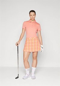 Golffelszerelés világos rózsaszín galléros pólóval és mintás szoknyanadrággal, narancs és sárga színekben. Fehér zokni és cipő szürke részletekkel, golfütőt tartva.