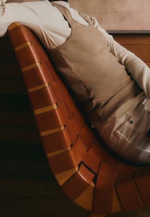 Person lehnt sich auf einem geflochtenen Lederstuhl zurück und trägt einen beigen ärmellosen Pullover über einem weißen Langarmhemd und hellen Hosen.