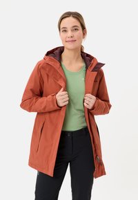 Chaqueta impermeable de color óxido con capucha, forro contrastado, bolsillos con cremallera y puños ajustables, llevada sobre una camiseta verde.
