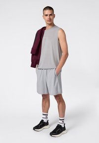 Uomo che indossa una maglietta senza maniche grigia e pantaloni corti, con una giacca bordeaux appoggiata sulla spalla, in piedi con le mani nelle tasche, indossando scarpe da ginnastica nere.