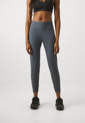 Femme portant un pantalon de sport fuselé gris foncé, un soutien-gorge de sport noir et des baskets noires, debout devant un fond clair uni.