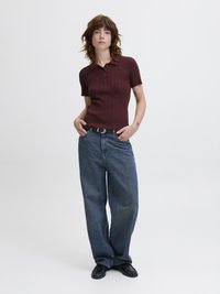 Kvinne med kort bølget hår iført en maroon kortermet ribbestrikket polo, løse blå jeans, svart belte og svarte sko, stående med hendene i lommene.