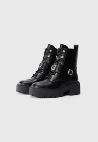 Stivali alla caviglia in pelle nera con punte arrotondate, lacci, fibbie metalliche e una suola spessa con un design a motivi.