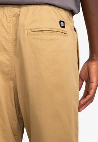 Khaki bukser med en afslappet pasform, der har en baglomme og et mærket sort label. Stoffet er glat med en let tekstur.