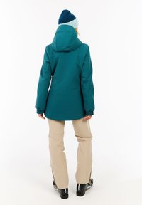 Türkisfarbene wasserdichte Jacke mit Kapuze, elastischen Bündchen und lockerer Passform, kombiniert mit beigen Hosen und schwarzen Skischuhen.