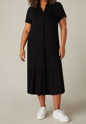 Frau trägt ein schwarzes, kurzärmeliges Midi-Kleid mit Knopfleiste und gerafftem Saum sowie weiße Plateau-Sneaker und steht vor einem schlichten Hintergrund.