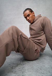 Bruine hoodie en trainingsbroek gemaakt van zacht materiaal, met een rits aan de voorkant, zijzakken en een pluche accent op de broek.