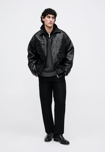 Giovane uomo con capelli ricci che indossa una giacca di pelle nera, un maglione grigio scuro, pantaloni neri e scarpe eleganti nere, in piedi su uno sfondo bianco.
