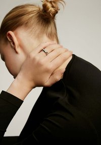 Anillo de plata en una mano que reposa sobre un hombro, con una blusa de manga larga negra. Cabello recogido en un moño ordenado, fondo neutro.