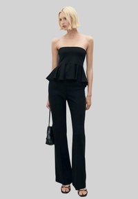 Čierny bezramenný peplum top v kombinácii s priliehajúcimi čiernymi nohavicami; modelka drží malú čiernu tašku, ktorá vystihuje hladkú textúru a moderný siluetu.