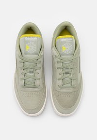 Gröna mockasneakers med en slät yta, med en perforerad tåbox, kontrasterande gula detaljer och en klassisk snörning.
