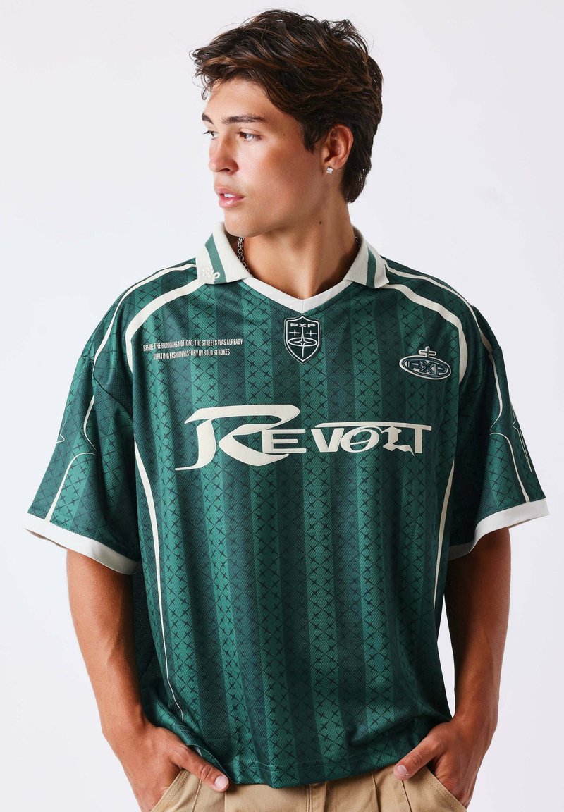 Jeune homme aux cheveux bruns courts portant un maillot de sport vert "Revolt", regardant vers la gauche avec les mains dans les poches d’un pantalon beige.
